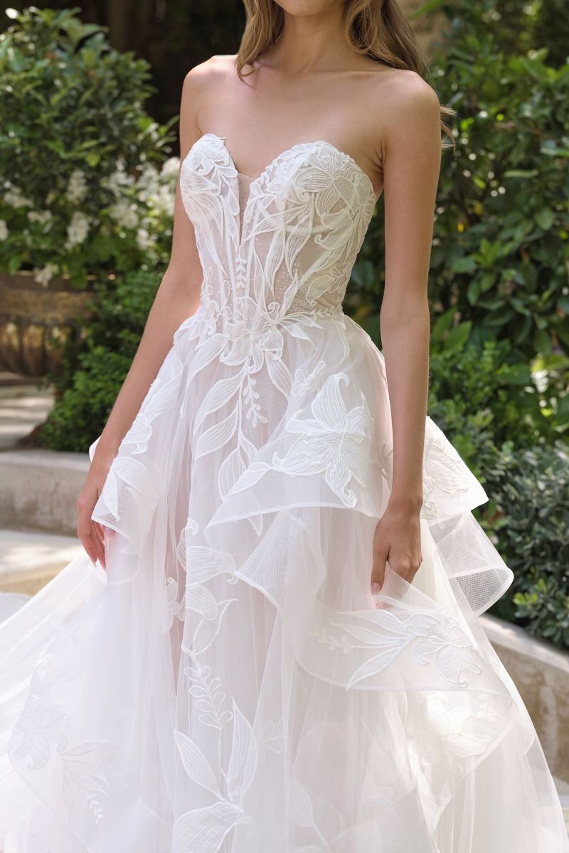 Vestido de novia que presenta un escote corazón sin tirantes y un corpiño semitransparente con varillas y una falda de tul escalonada en cascada está adornada con un delicado diseño floral de lirios estrellados
